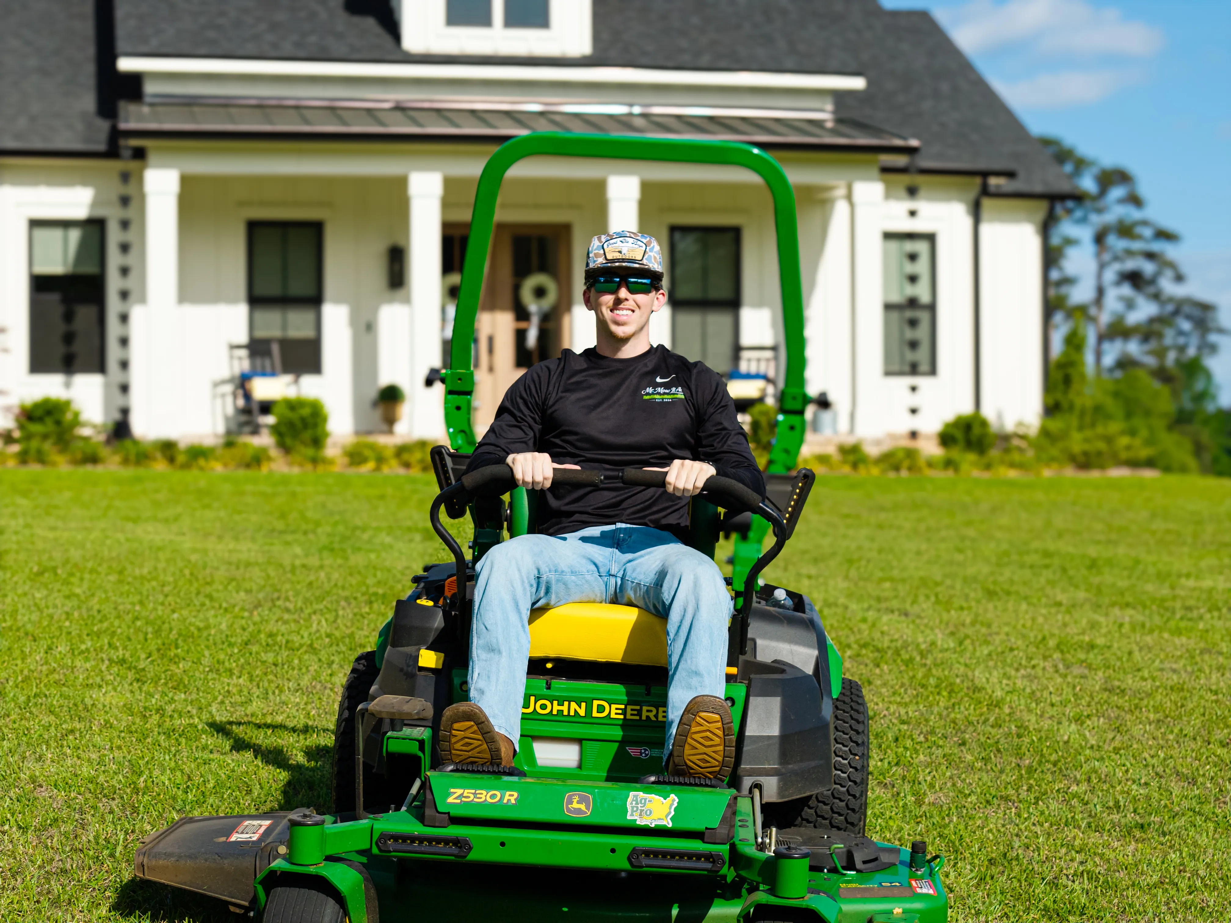 Mr. MowItAll mowing a manicured Tallahassee lawn on a green John Deere zero-turn mower in front of a modern farmhouse