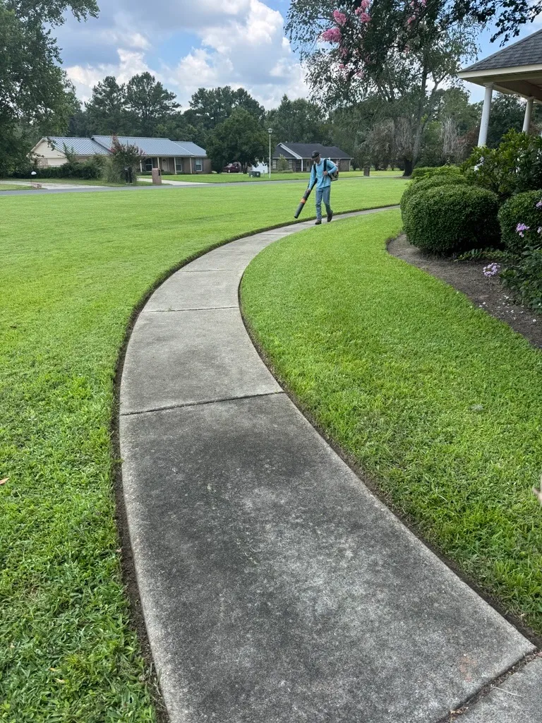 Mr. MowItAll crew member using a backpack blower to clear clippings off a curving sidewalk beside a freshly cut lawn